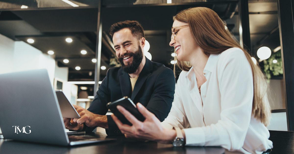 Two team members discussing SEO strategies and improvements for the business while smiling and working on their laptops and mobile devices in a modern office setting.