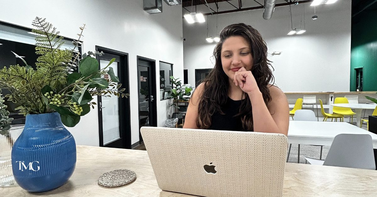 Giselle, Senior Designer at TMG Marketing Partners, works thoughtfully on her laptop in a modern, open office space—capturing the spirit of a brand refresh with purpose. A stylish blue vase with greenery adds a creative touch to the workspace.