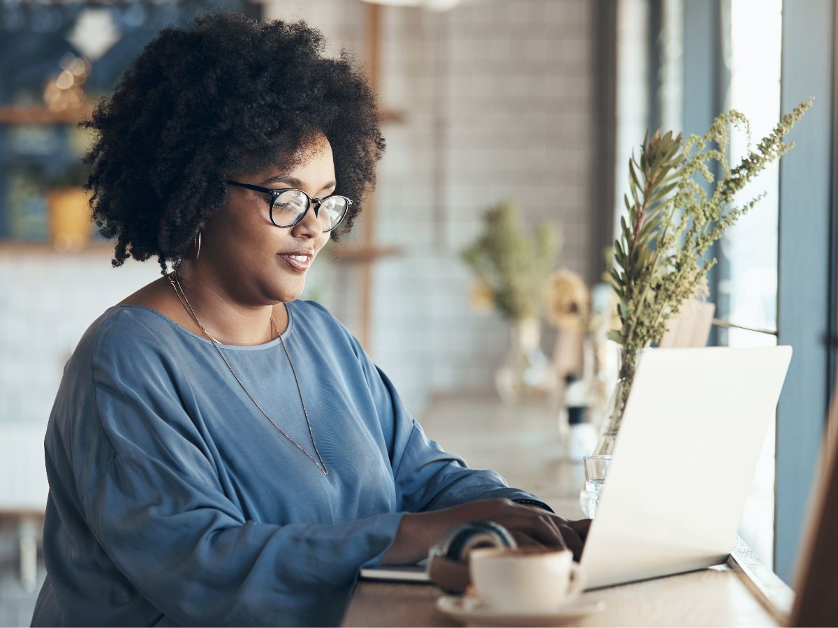 A woman working on a laptop in a bright café setting with a coffee cup and greenery on the table. The image conveys a professional and inviting atmosphere, encouraging viewers to book a consult.