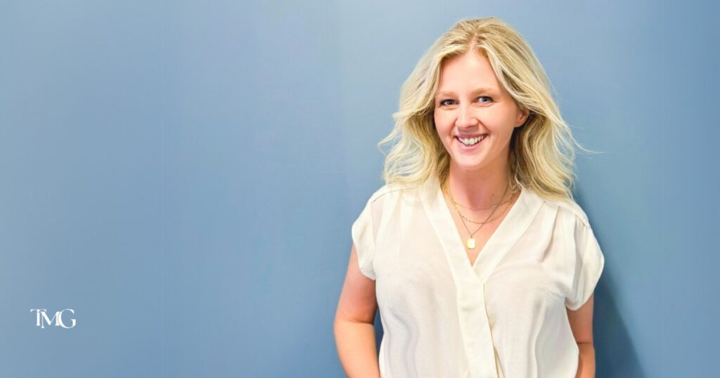 Cristalle Gleason, now 100% owner of TMG Marketing Partners, stands against a light blue background, wearing a white blouse and layered necklaces, exuding a professional yet approachable demeanor. The logo for TMG Marketing Partners is displayed in the corner, representing a Christian-led business focused on values-driven marketing solutions.