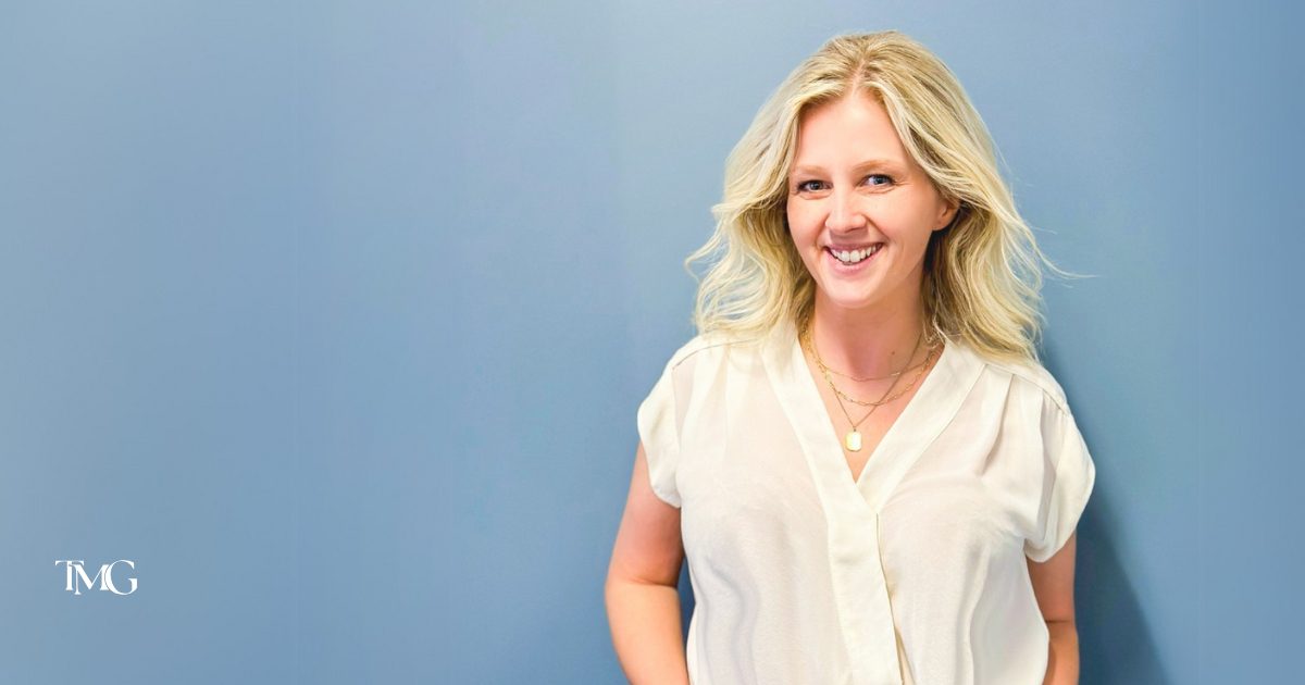 Cristalle Gleason, now 100% owner of TMG Marketing Partners, stands against a light blue background, wearing a white blouse and layered necklaces, exuding a professional yet approachable demeanor. The logo for TMG Marketing Partners is displayed in the corner, representing a Christian-led business focused on values-driven marketing solutions.