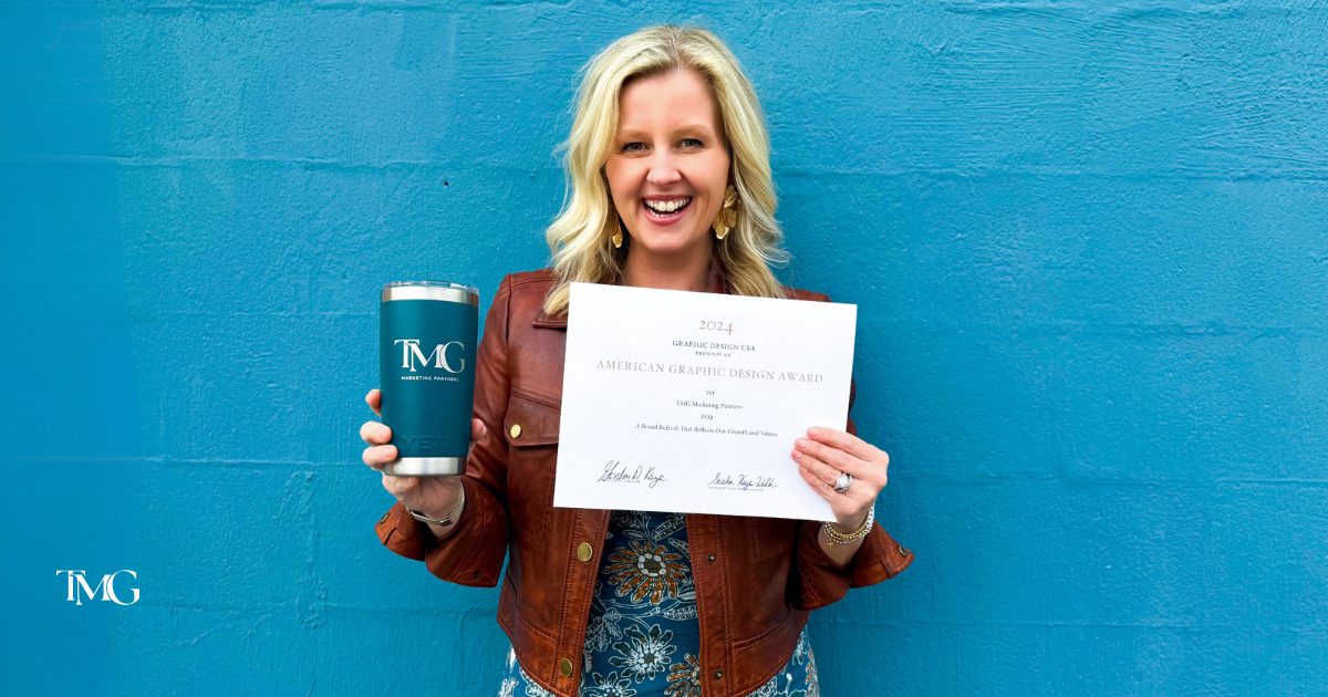 The TMG Marketing Partner Owner, Cristalle, wearing a brown leather jacket and a floral dress, smiles while holding a Graphic Design Award certificate in one hand and a branded award-winning logo tumbler in the other. The certificate, awarded by Graphic Design USA in 2024, recognizes TMG Marketing Partners for outstanding branding. She stands against a vibrant blue wall with the TMG logo subtly placed in the background.
