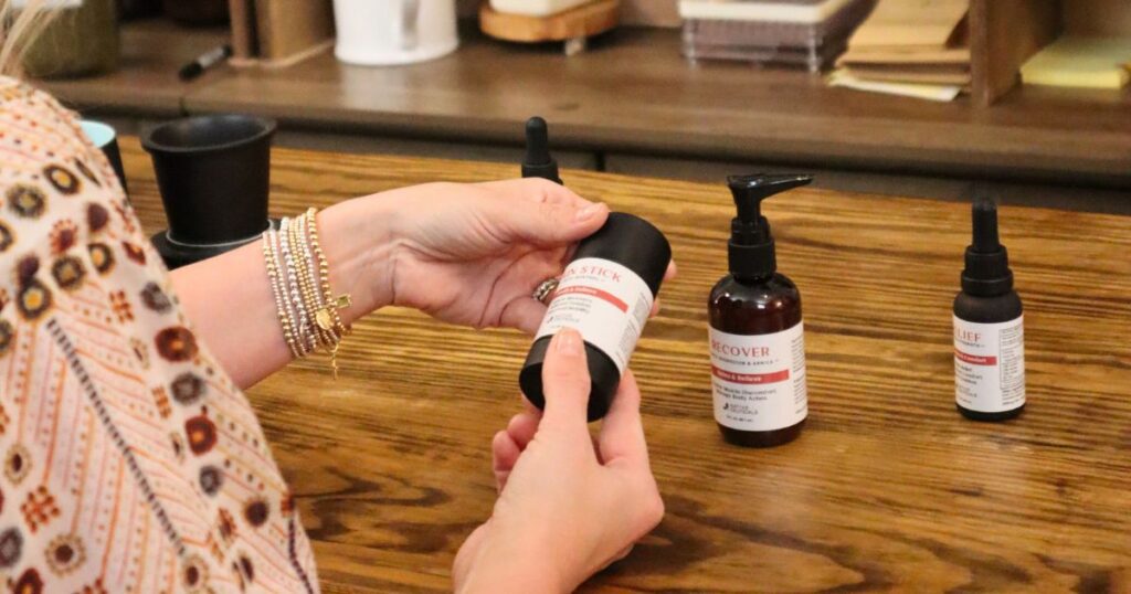 Hands holding redesigned Native Ceuticals pain stick while browsing companion wellness products like Recover and Relief on display.