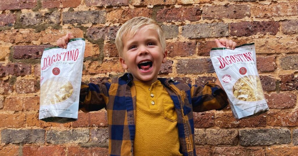 Young boy holding Dagostino Pasta bags against a rustic brick wall showing excitement for handmade pasta