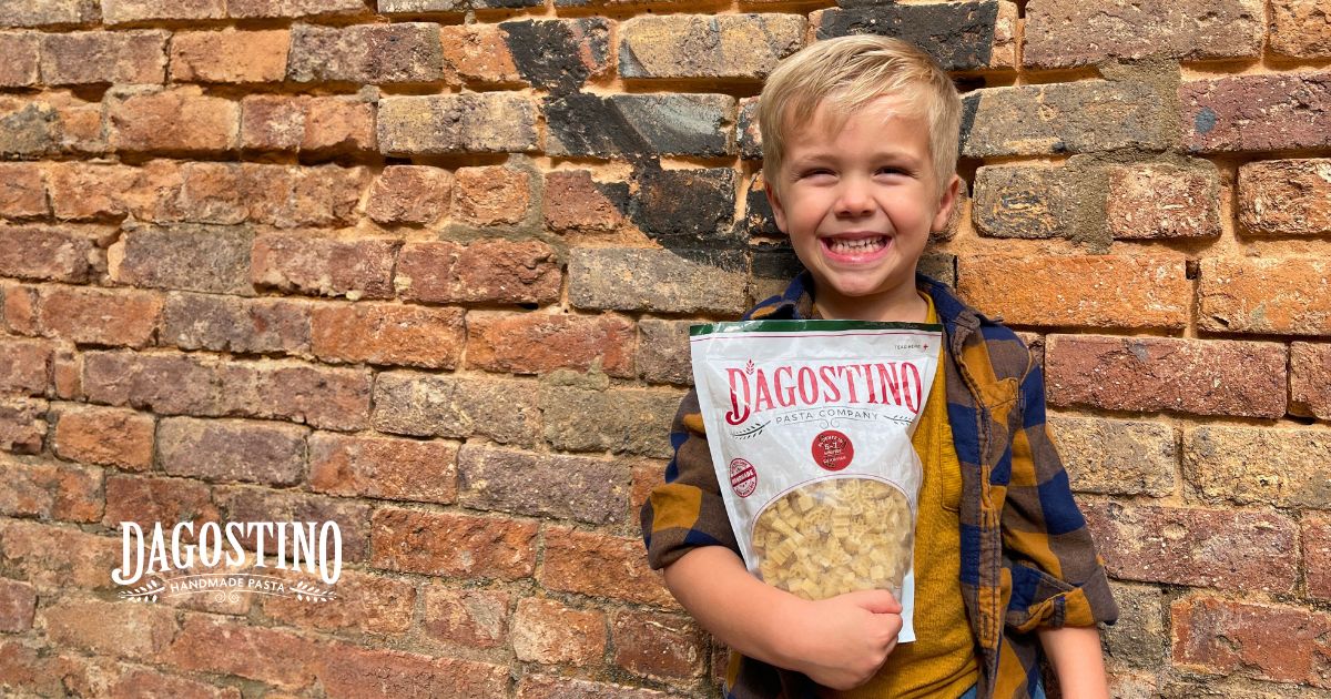 Happy child holding a bag of Dagostino Pasta against a vintage brick wall, representing family appeal