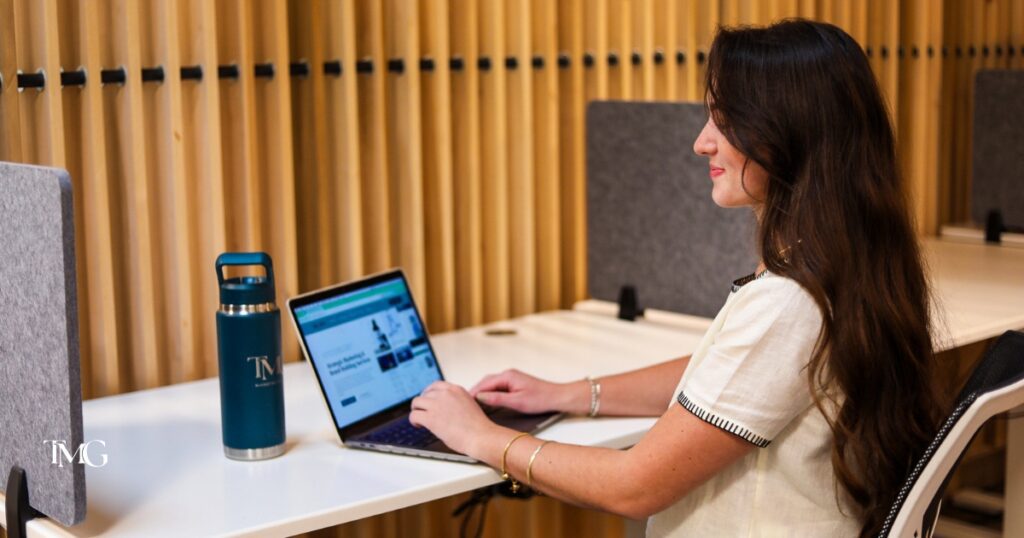 A woman working at a laptop in a modern office setting, with the TMG logo on her water bottle, representing the search for great keywords for online visibility and future proofing for long-term business growth.