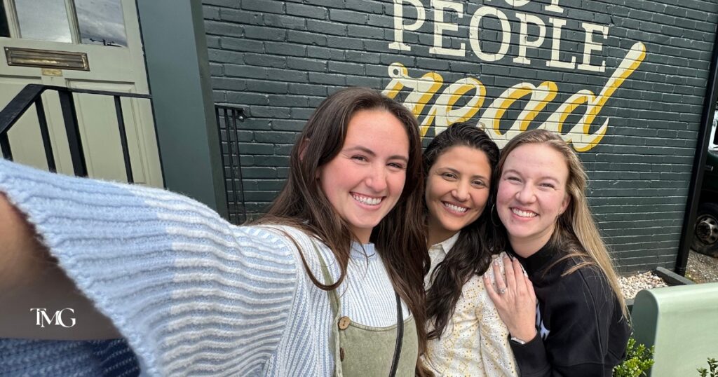 Three smiling team members from TMG Marketing Partners pose in front of a mural that reads "People read," reflecting the company’s focus on authentic branding and brand authenticity through real connection.