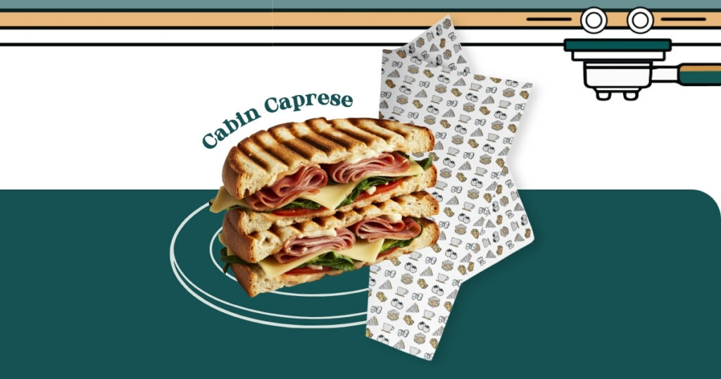 Griddled Cabin Caprese panini with ham, cheese, tomato, and greens on toasted bread, displayed on branded patterned wrapper