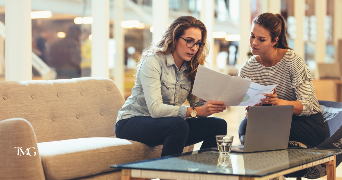 Two women collaborating on a digital marketing audit, reviewing printed reports on a couch with a laptop open on the coffee table.