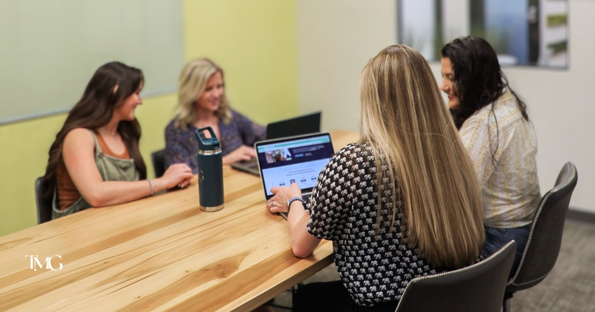 Four women gathered around a wooden conference table with laptops, discussing marketing trends and content marketing strategy for their team.