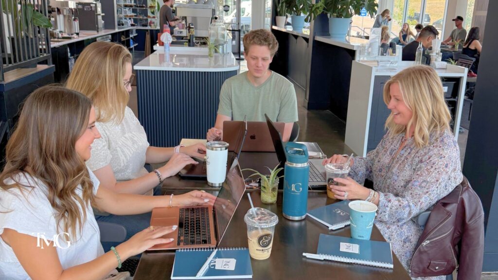 TMG team members work on laptops around a café table with coffee and branded notebooks, mapping out how AI in marketing fits a digital strategy.