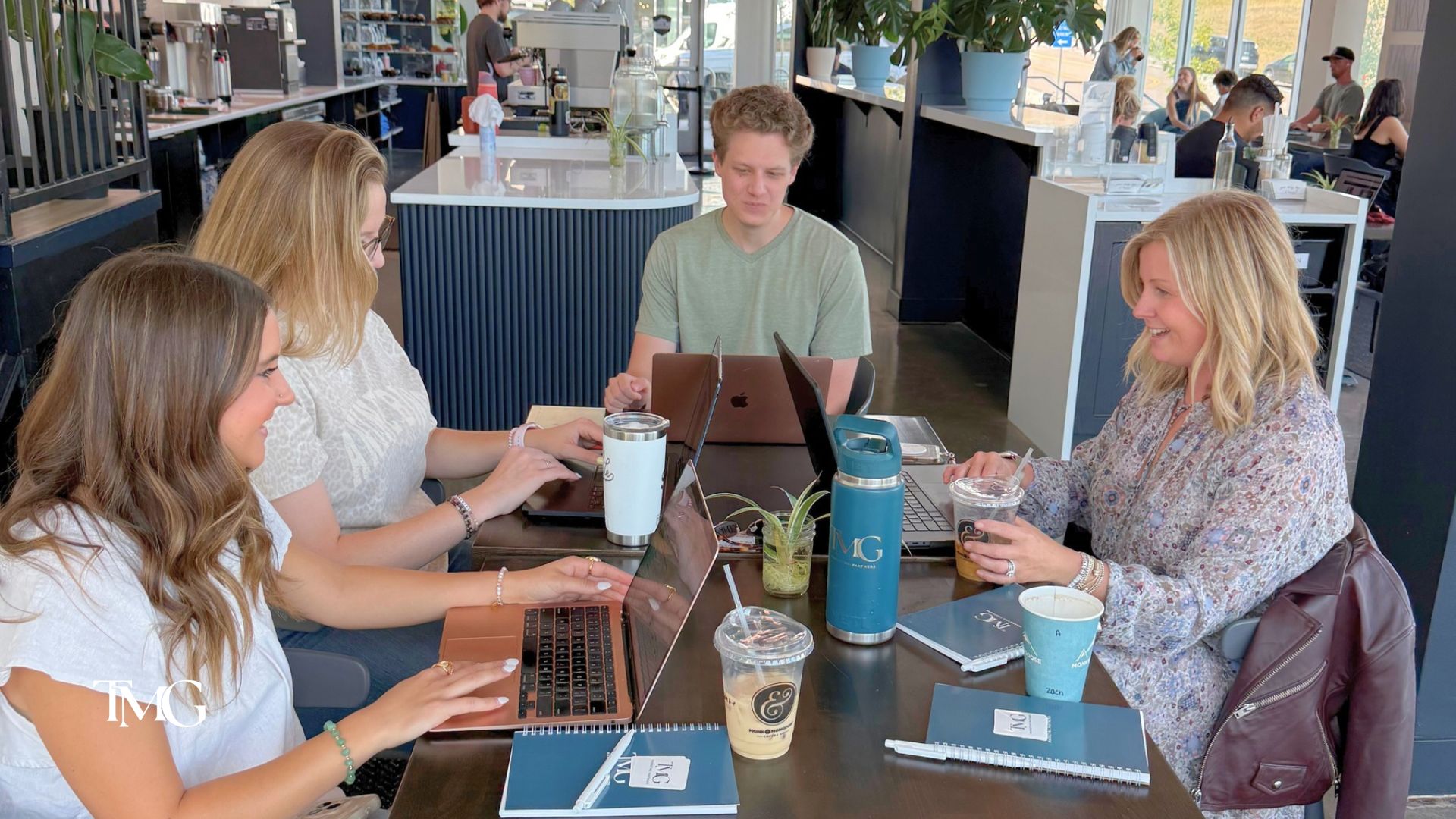 TMG team members work on laptops around a café table with coffee and branded notebooks, mapping out how AI in marketing fits a digital strategy.